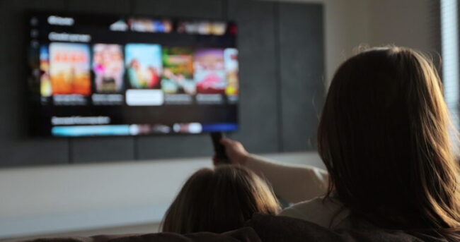 Woman selects tv channels with remote control at living room on sofa. Woman with her little daughter controls TV using a modern remote control.