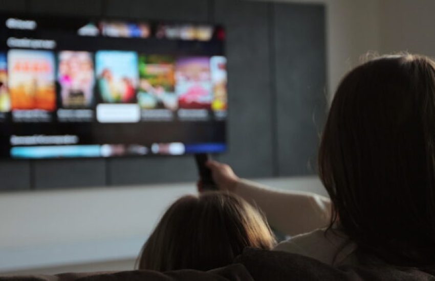 Woman selects tv channels with remote control at living room on sofa. Woman with her little daughter controls TV using a modern remote control